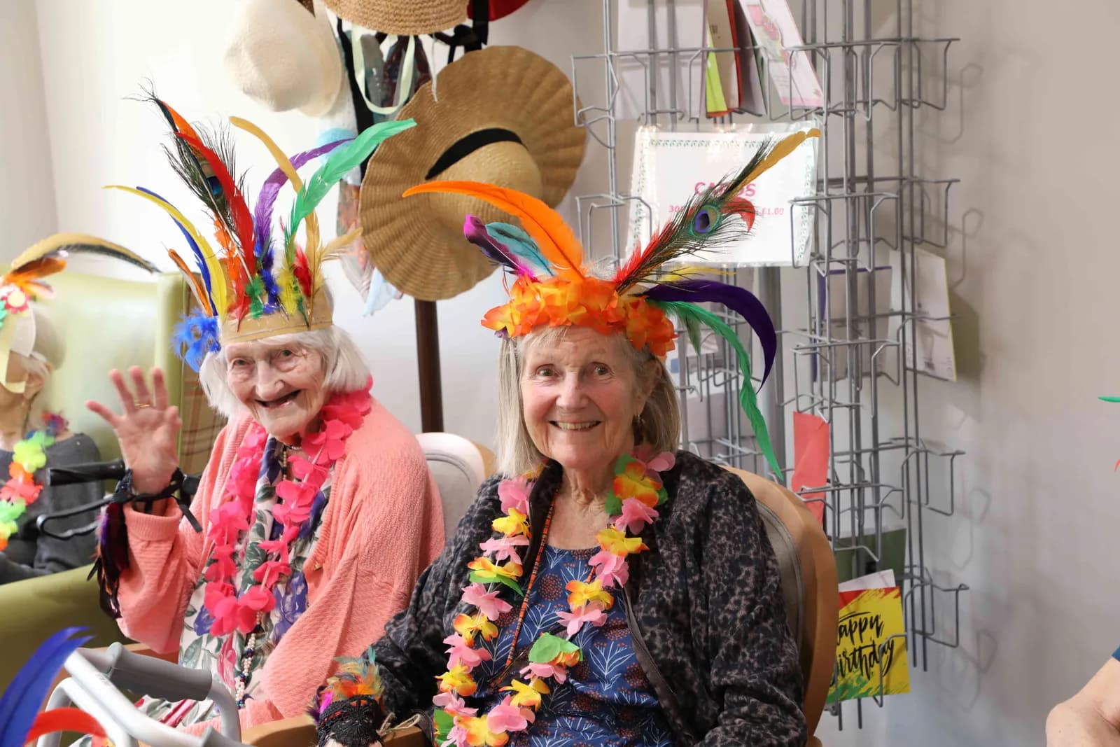 Two smiling elderly women wear brightly colored feather headpieces and leis, with one woman waving at the camera.