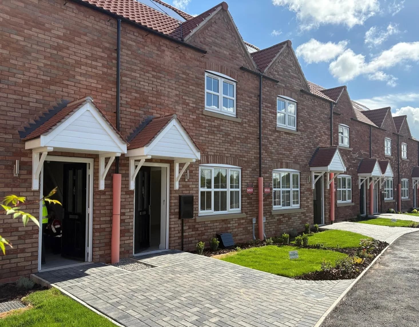A row of newly built red brick townhouses with white framed windows, dark brown doors, and small white canopies over each entrance, with paved driveways and small patches of green lawn in front of them.