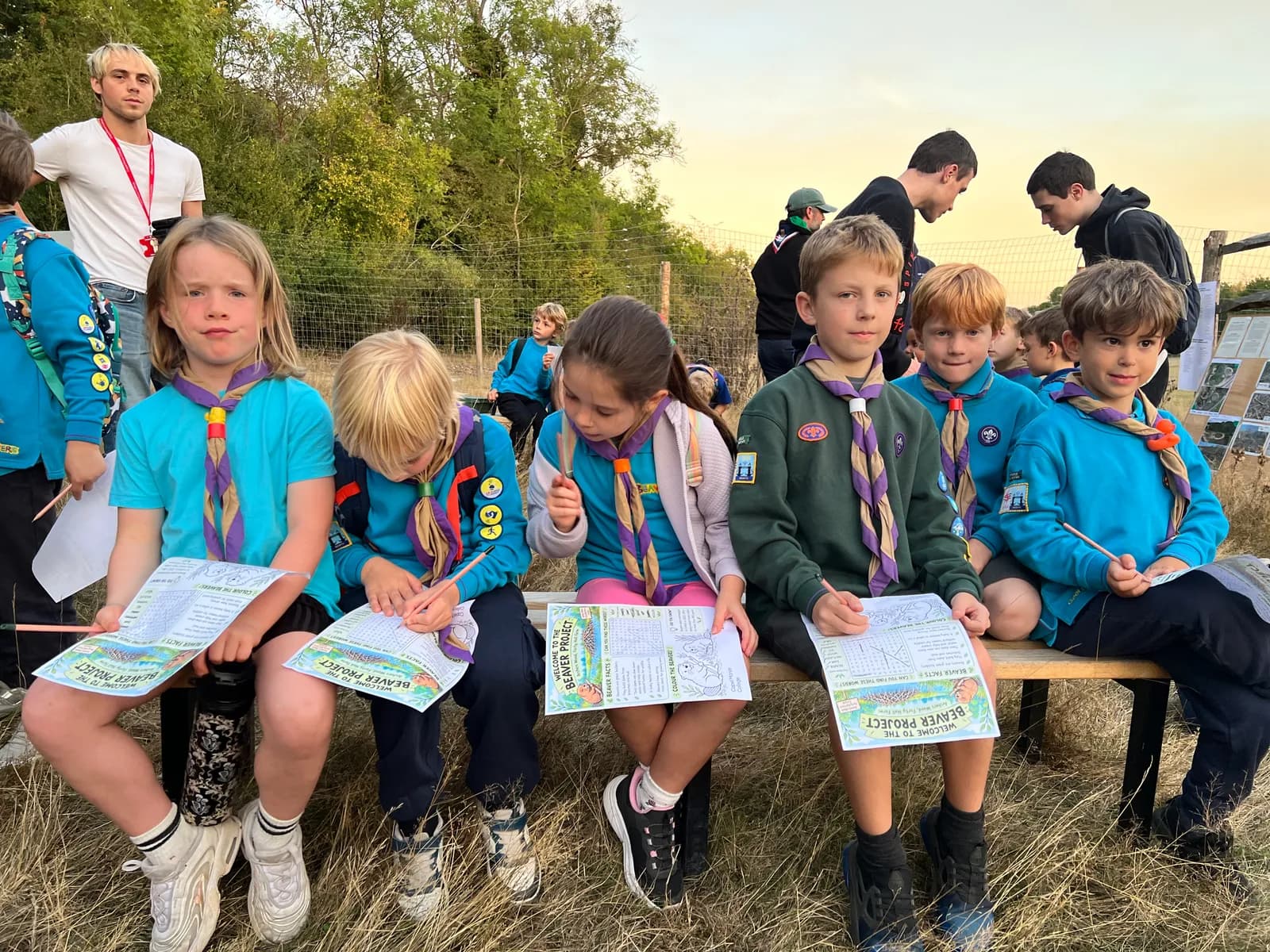 A group of young children wearing scout uniforms and holding papers sit on a bench outdoors, with adults standing in the background.