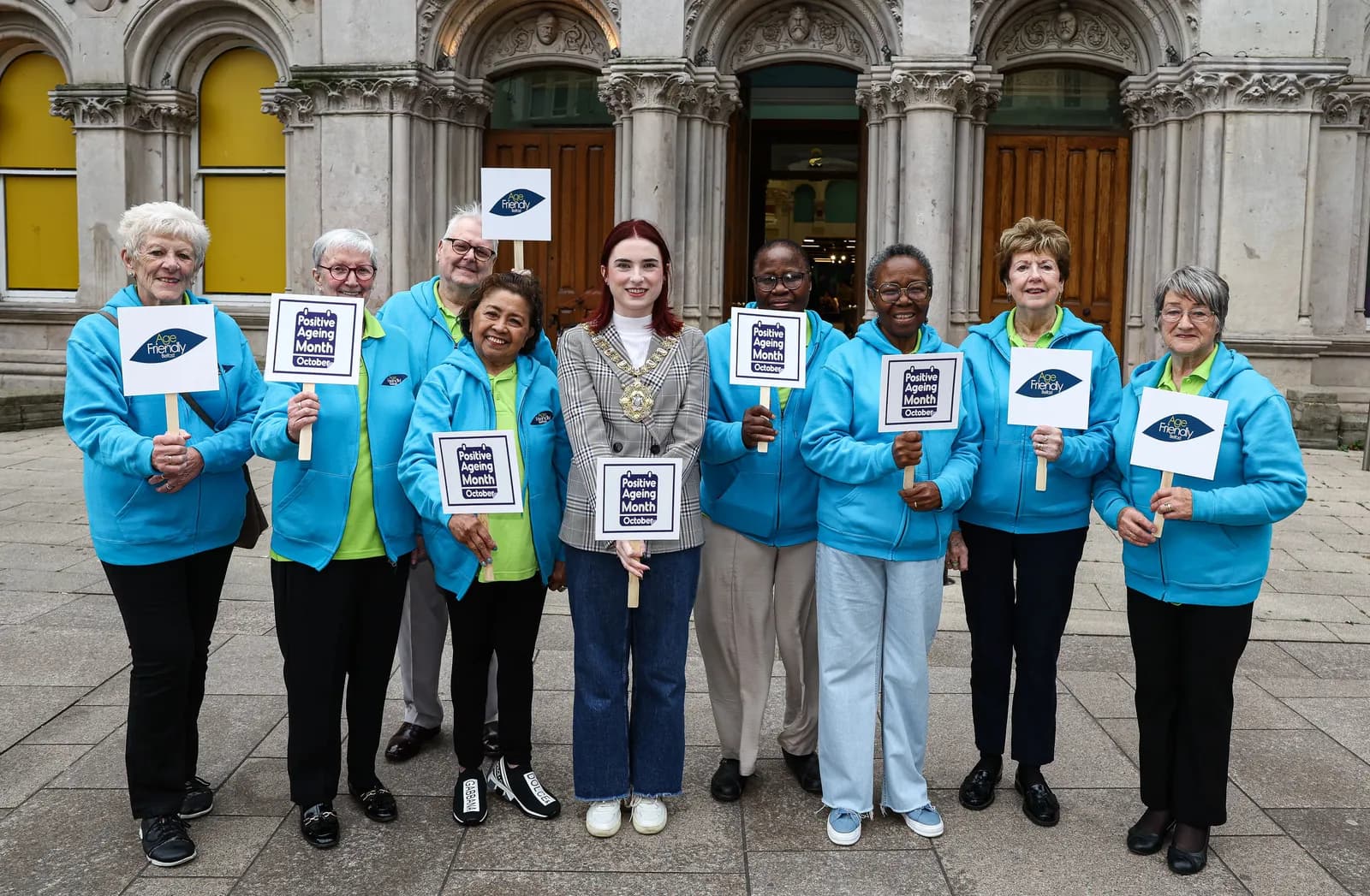 A group of older adults and one younger woman in a gray checkered blazer stand in front of a stone building holding signs that say "Age Friendly" or "Positive Ageing Month."