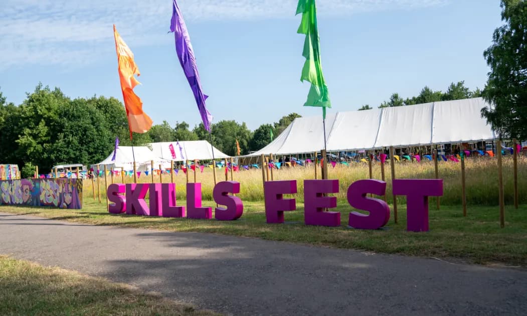 Large purple letters spelling "SKILLS FEST" sit on a grassy field with white tents and colorful flags in the background under a blue sky.