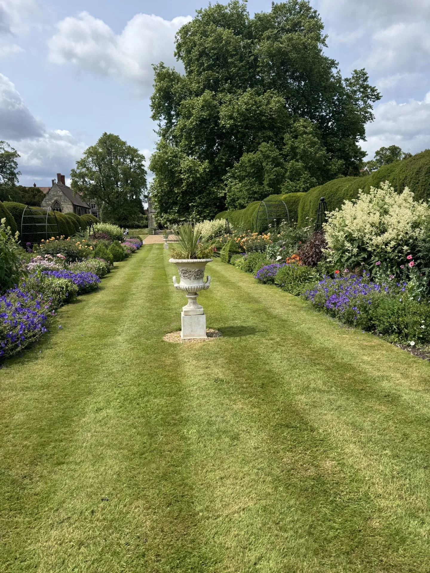 A grand garden path lined with vibrant flowers and tall hedges, leading to an ornate stone urn and distant buildings under a partly cloudy sky.