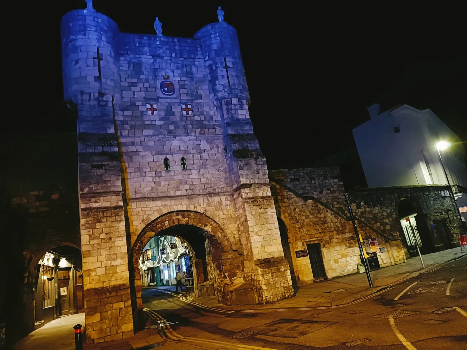 A large stone archway with two towers on top, illuminated with blue light, stands over a narrow street at night, with streetlights casting a warm glow on the surrounding buildings.