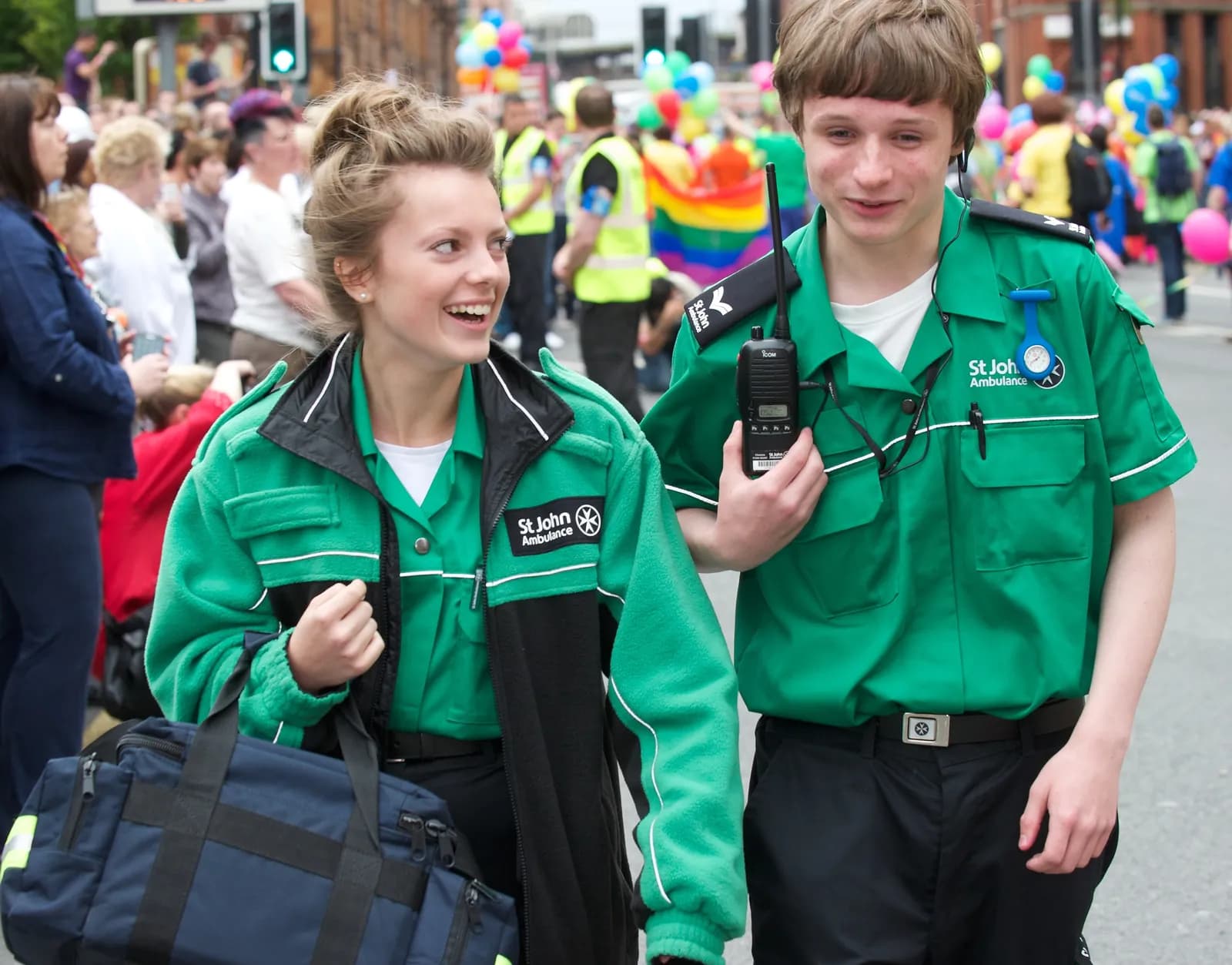 Two young St John Ambulance volunteers in green uniforms walk in a parade with crowds and balloons visible in the background.