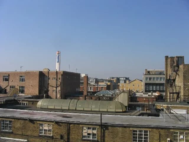 A wide view of urban rooftops stretching into the distance under a clear blue sky, featuring various brick buildings, chimneys, and a white tower with a blue logo.