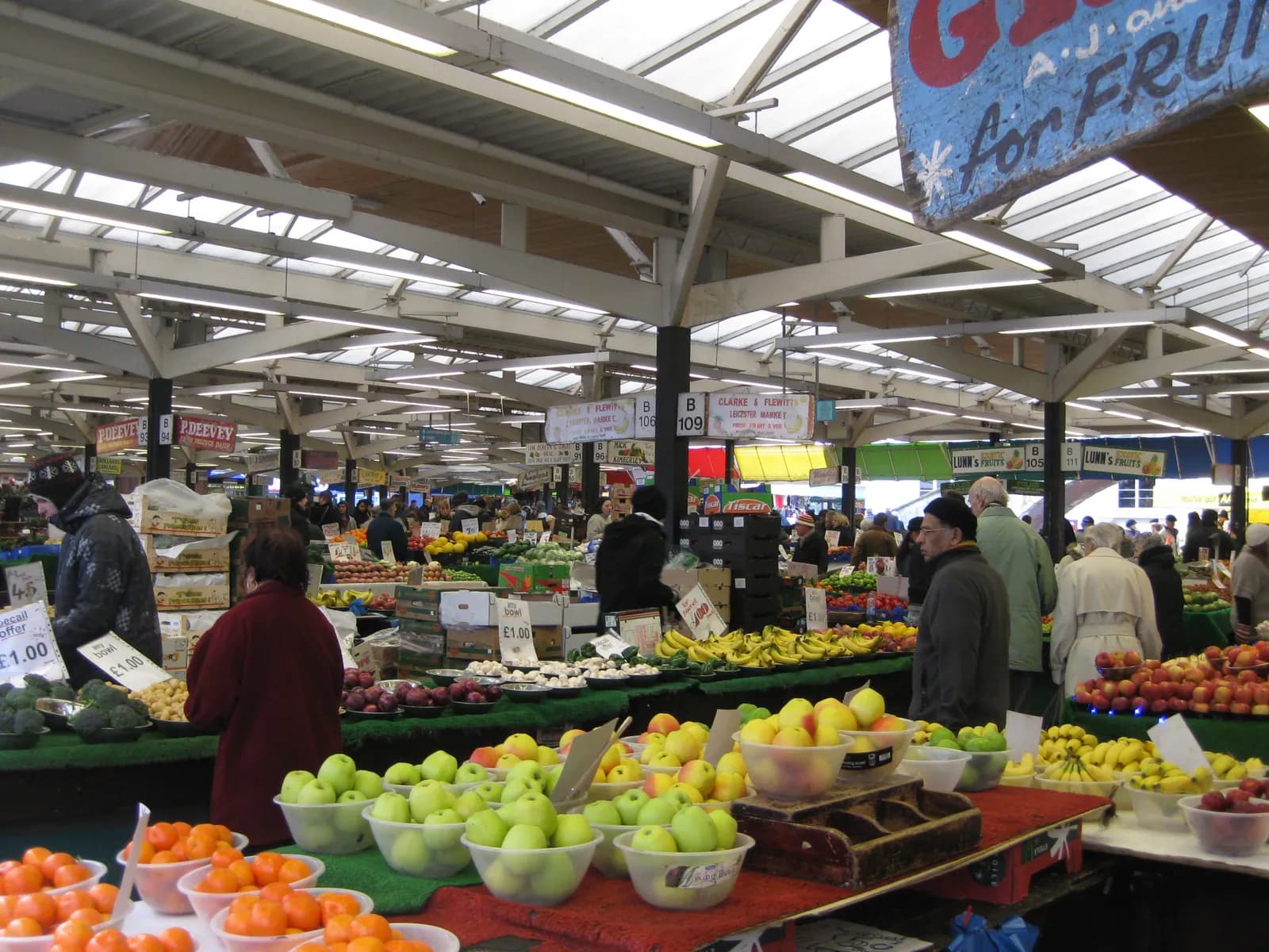 A bustling indoor market with people browsing various fruit and vegetable stalls under a translucent roof.