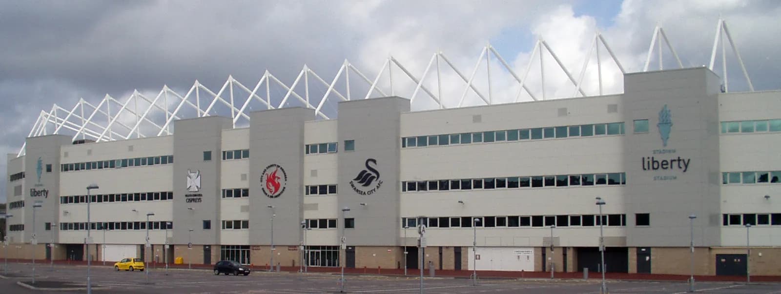 The white and grey exterior of Liberty Stadium with several logos visible on its facade and a partially cloudy sky overhead.