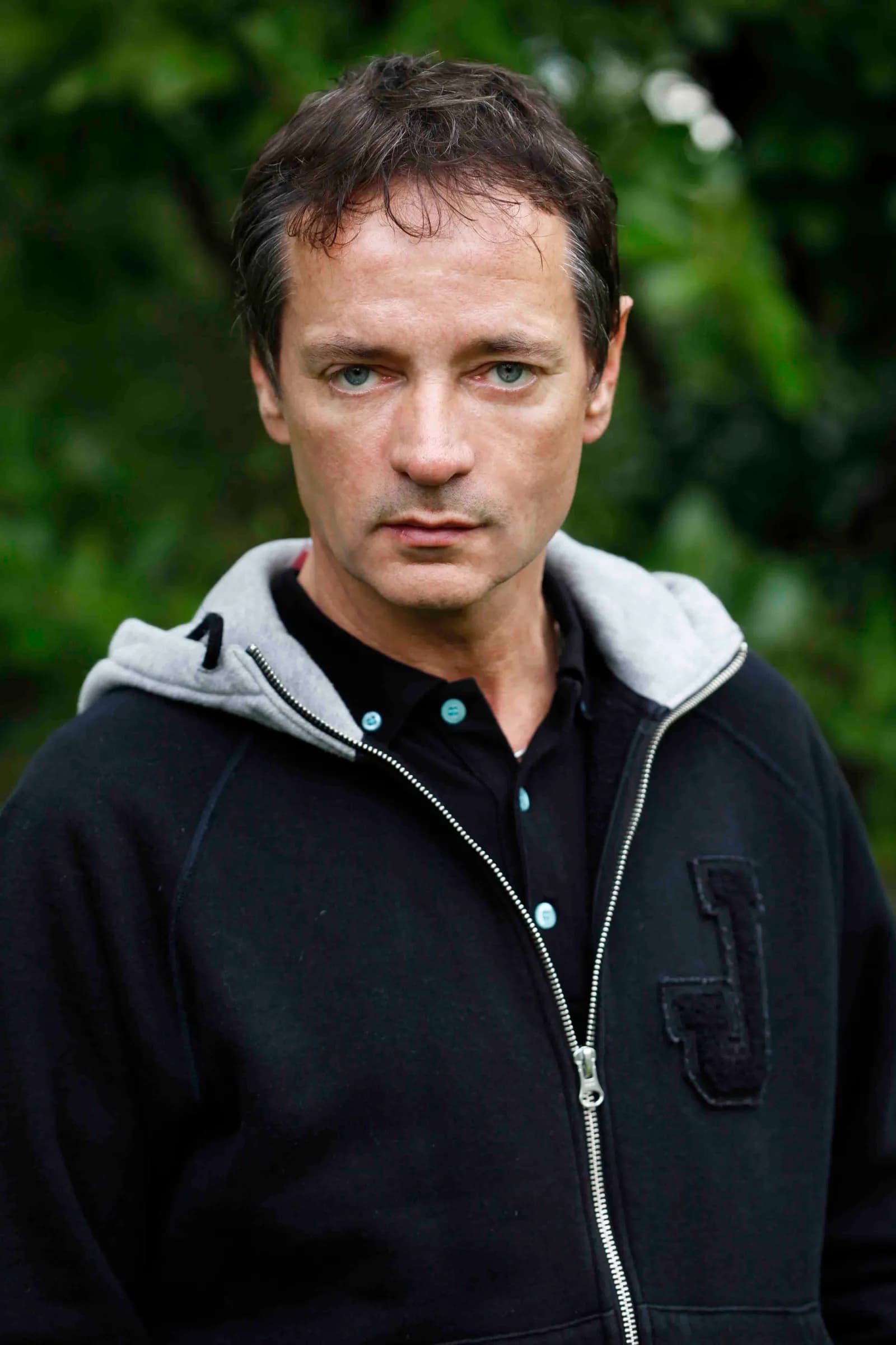 A man with dark hair and blue eyes wearing a black zipped hoodie over a black polo shirt with light blue buttons, standing outdoors with green foliage in the background.