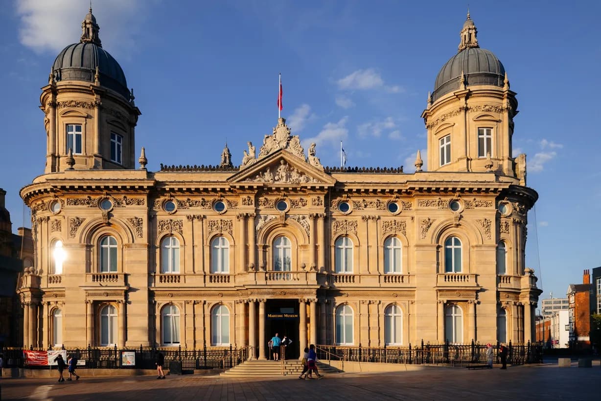 The historic Guildhall building stands under a clear blue sky, featuring intricate architectural details, domed towers, and a grand entrance, with people gathered on the steps and walking in the foreground.