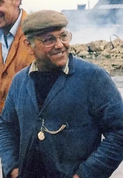 A man in glasses and a flat cap smiles, wearing a blue jacket with a pocket watch chained to the lapel.