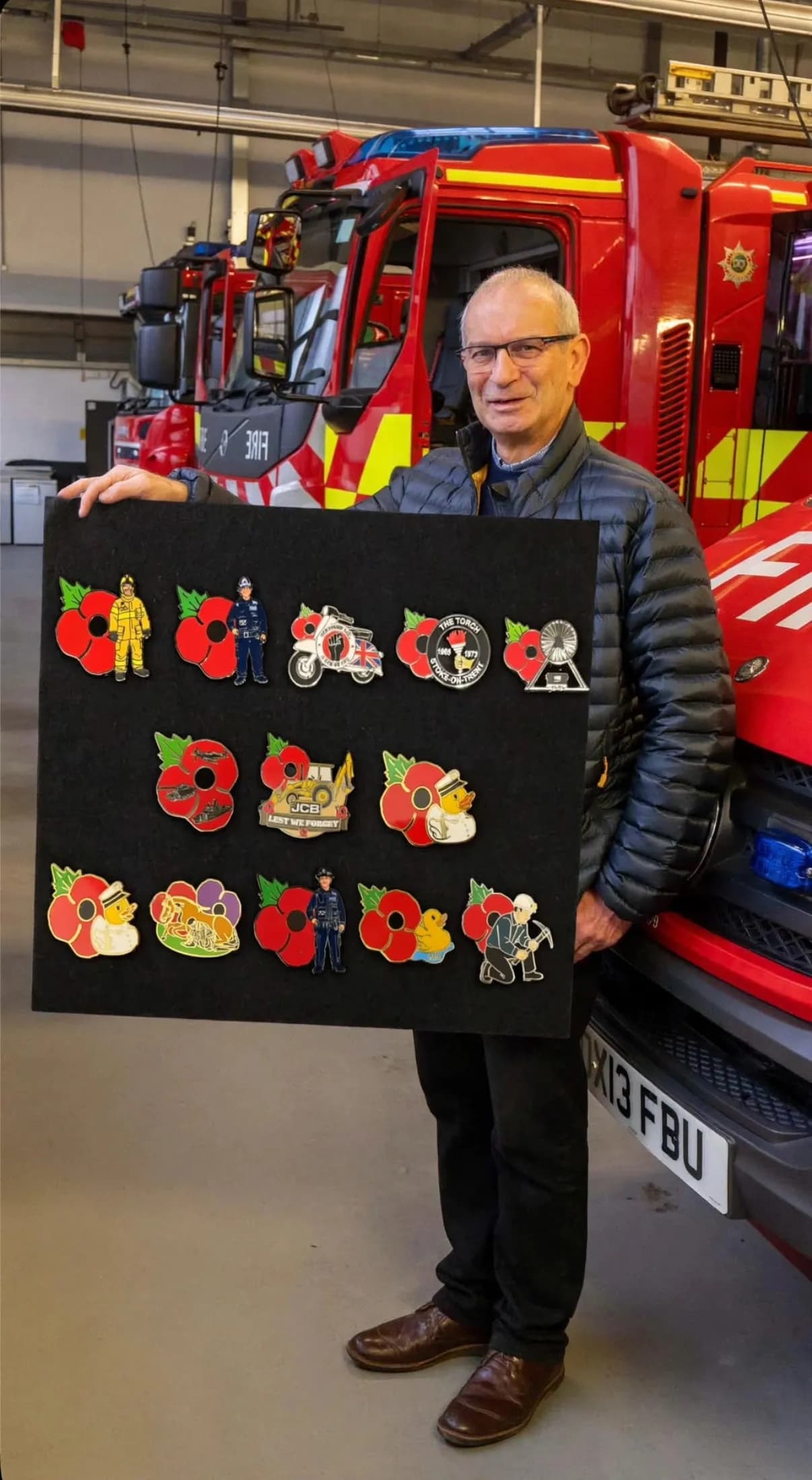 A man in glasses holds a black board covered in various poppy pins, standing in front of several red fire trucks.