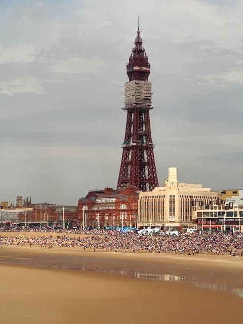 The Blackpool Tower stands tall on a cloudy day behind a crowded beach and buildings.