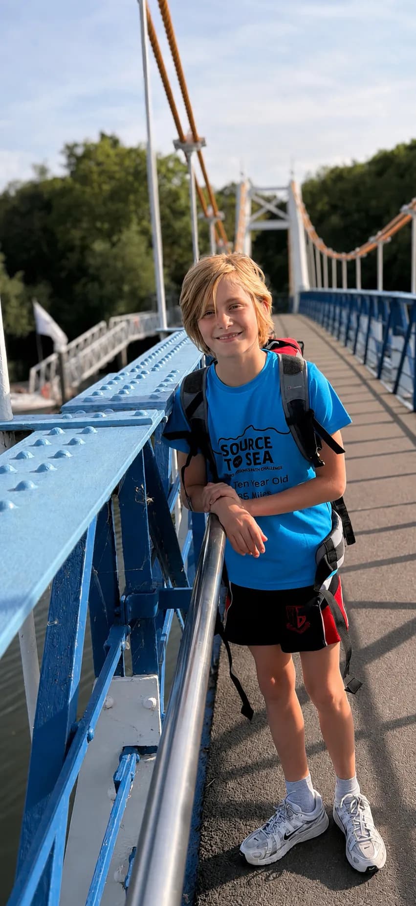 A smiling boy with blonde hair and a blue t-shirt leans on a blue bridge railing with green trees and blue sky in the background.