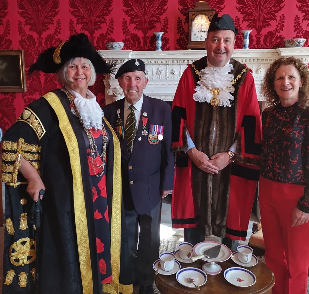 Four adults, two in ceremonial robes and a man in a military uniform, stand together in a room with a red patterned wallpaper and a fireplace.