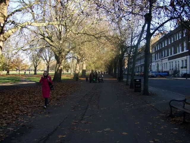 A woman in a red coat walks along a path covered in fallen leaves, with trees lining a park to her left and a street with buildings to her right.