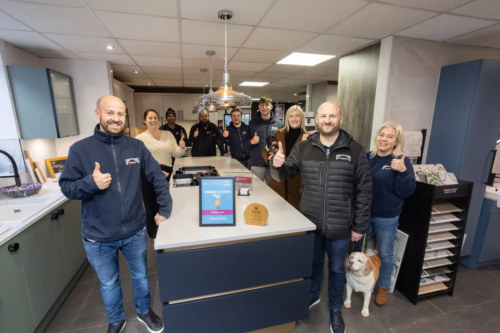 A team of eight people and one dog, all smiling and giving thumbs up, are gathered around a kitchen island in a showroom.