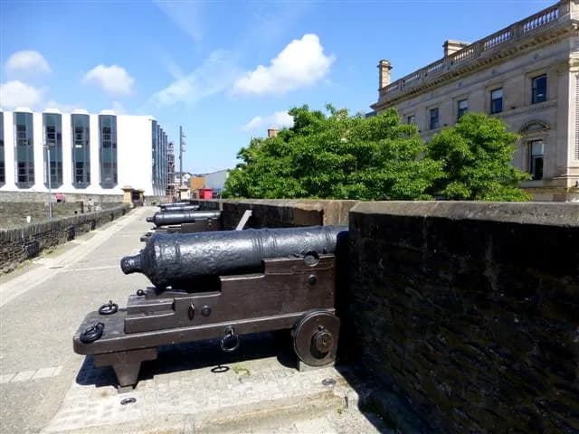 Several antique cannons lined up on a stone wall next to a modern white building and an older ornate stone building under a clear blue sky with fluffy white clouds.