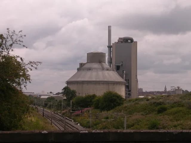 A large industrial facility with a round domed building and a tall rectangular structure with a smokestack under a cloudy sky.
