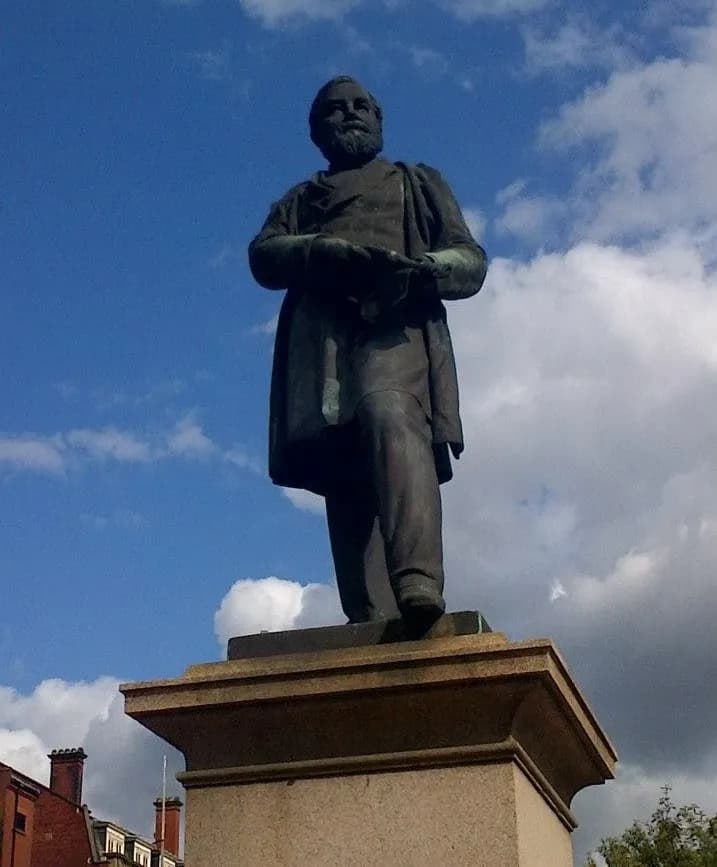 A bronze statue of a man with a beard, wearing a long coat, stands on a stone pedestal against a bright blue sky with white clouds.