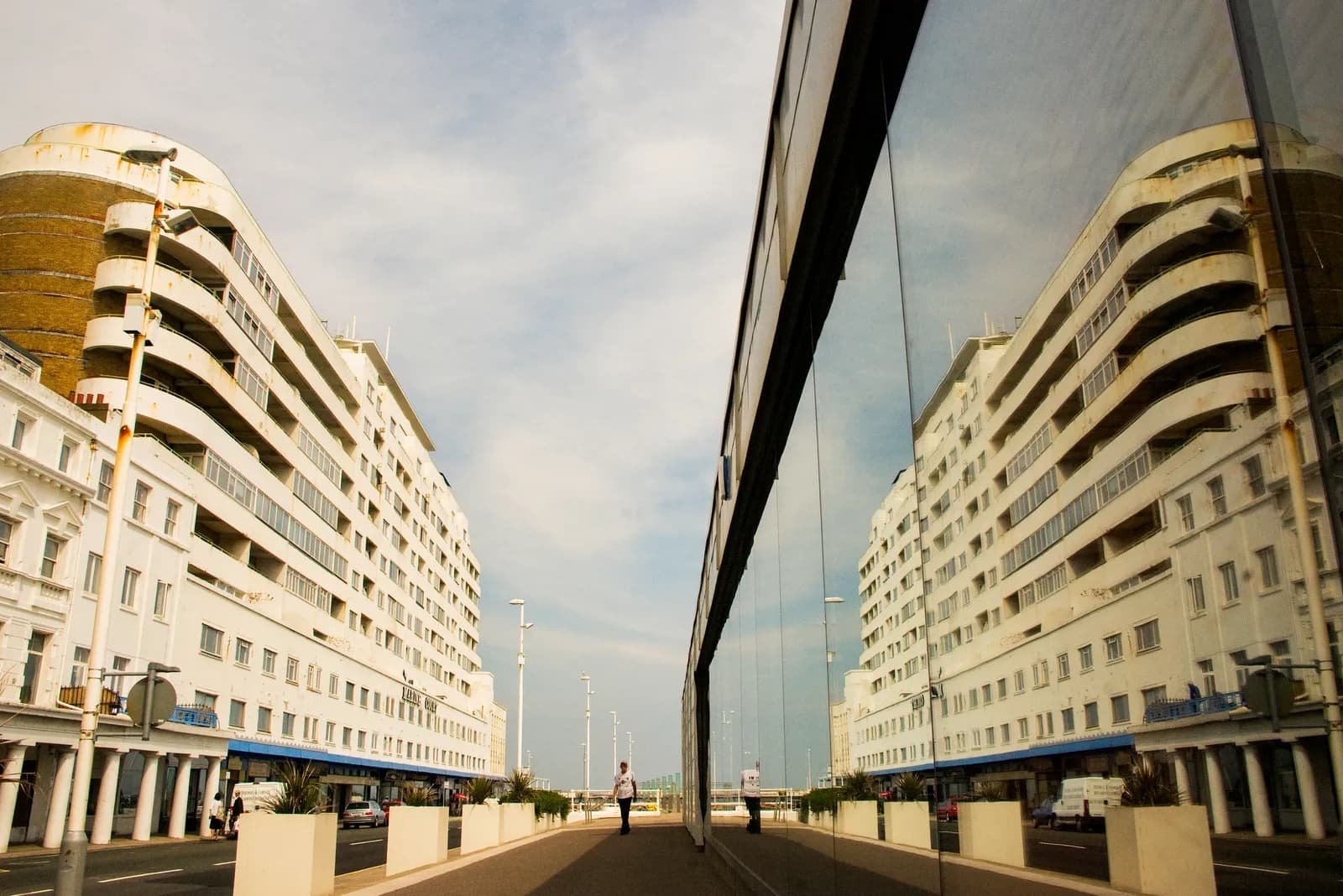 A large, curving white apartment building with balconies is reflected in the long glass wall of a building across the street.