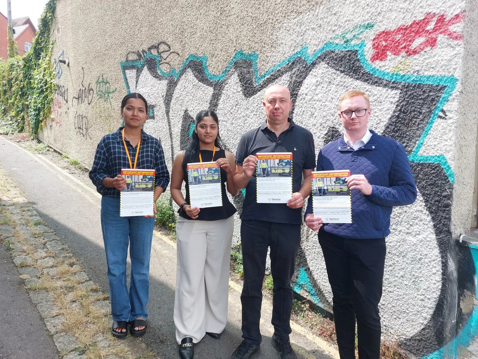 Four people, two women and two men, holding up flyers in front of a graffiti-covered wall, standing on a paved path.