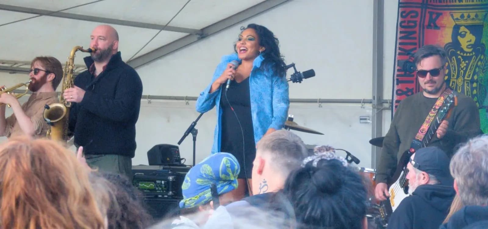 A band performing live on stage, with a female singer in a blue jacket holding a microphone and two male musicians playing saxophone and guitar.