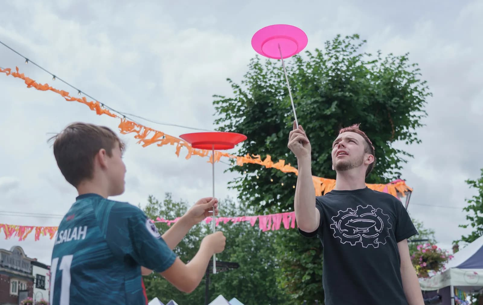 A young boy and an adult man are outside juggling spinning plates on sticks, with orange and pink streamers hanging in the background.