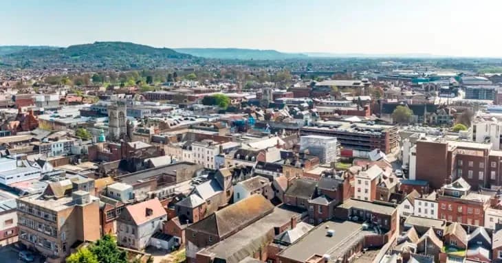 Elevated view of a town with various buildings, a church tower, and a distant forested hill under a clear sky.