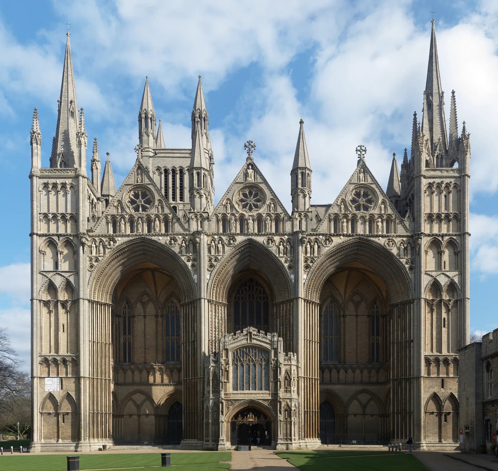 The ornate and imposing facade of Peterborough Cathedral is visible under a blue sky with scattered clouds, showcasing its intricate arched doorways, spires, and circular windows, with green grass in the foreground.