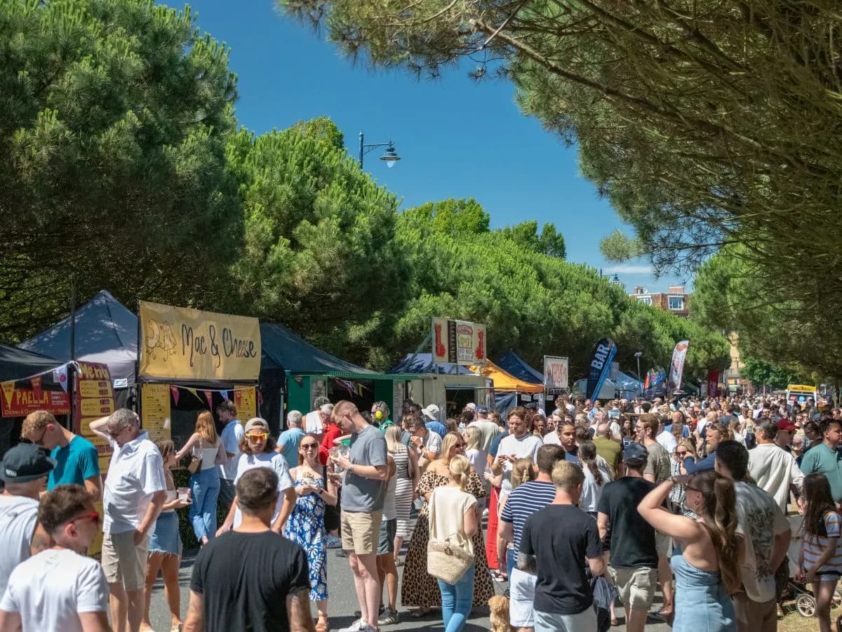 A crowded outdoor market scene with numerous people walking between food stalls under a clear blue sky and lush green trees.