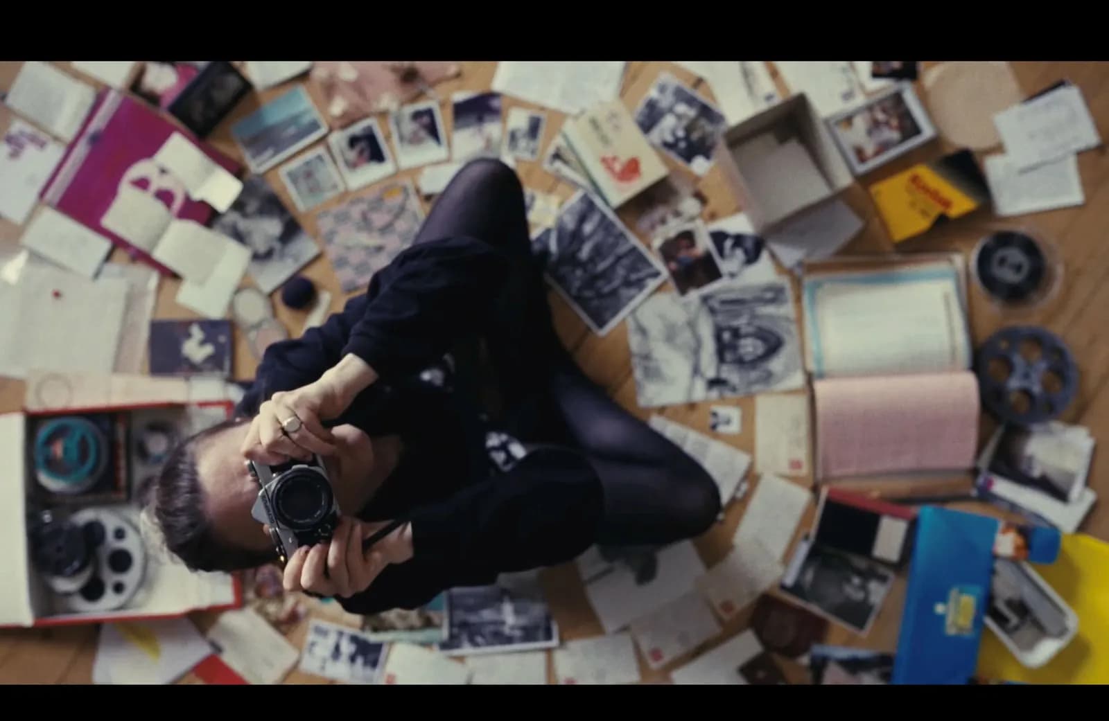 A person sitting on a wooden floor surrounded by an assortment of photographs, letters, and film reels, holding a camera to their eye as if taking a photo.