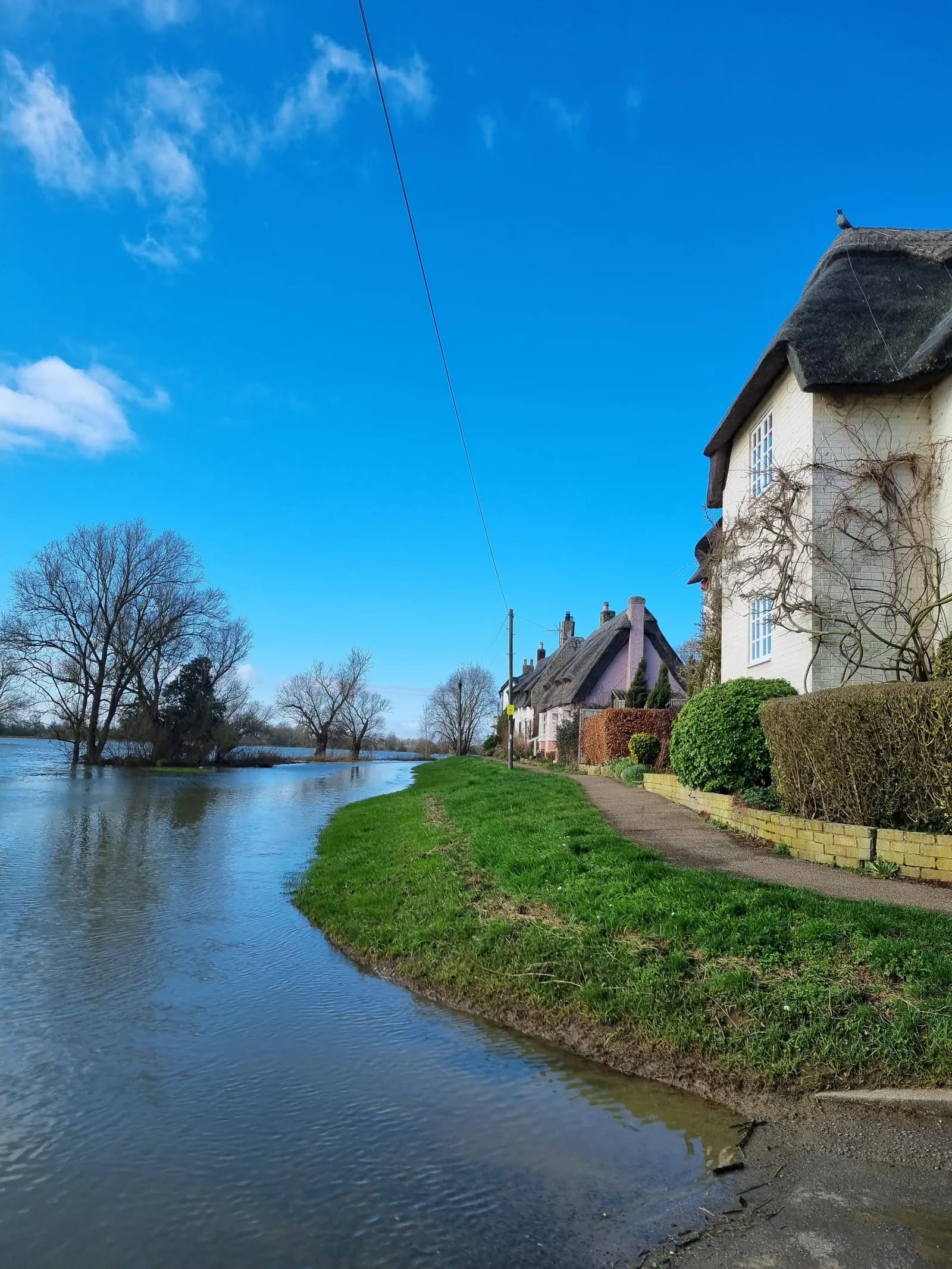 A flooded river with grassy banks, a path, and houses with thatched roofs under a bright blue sky.