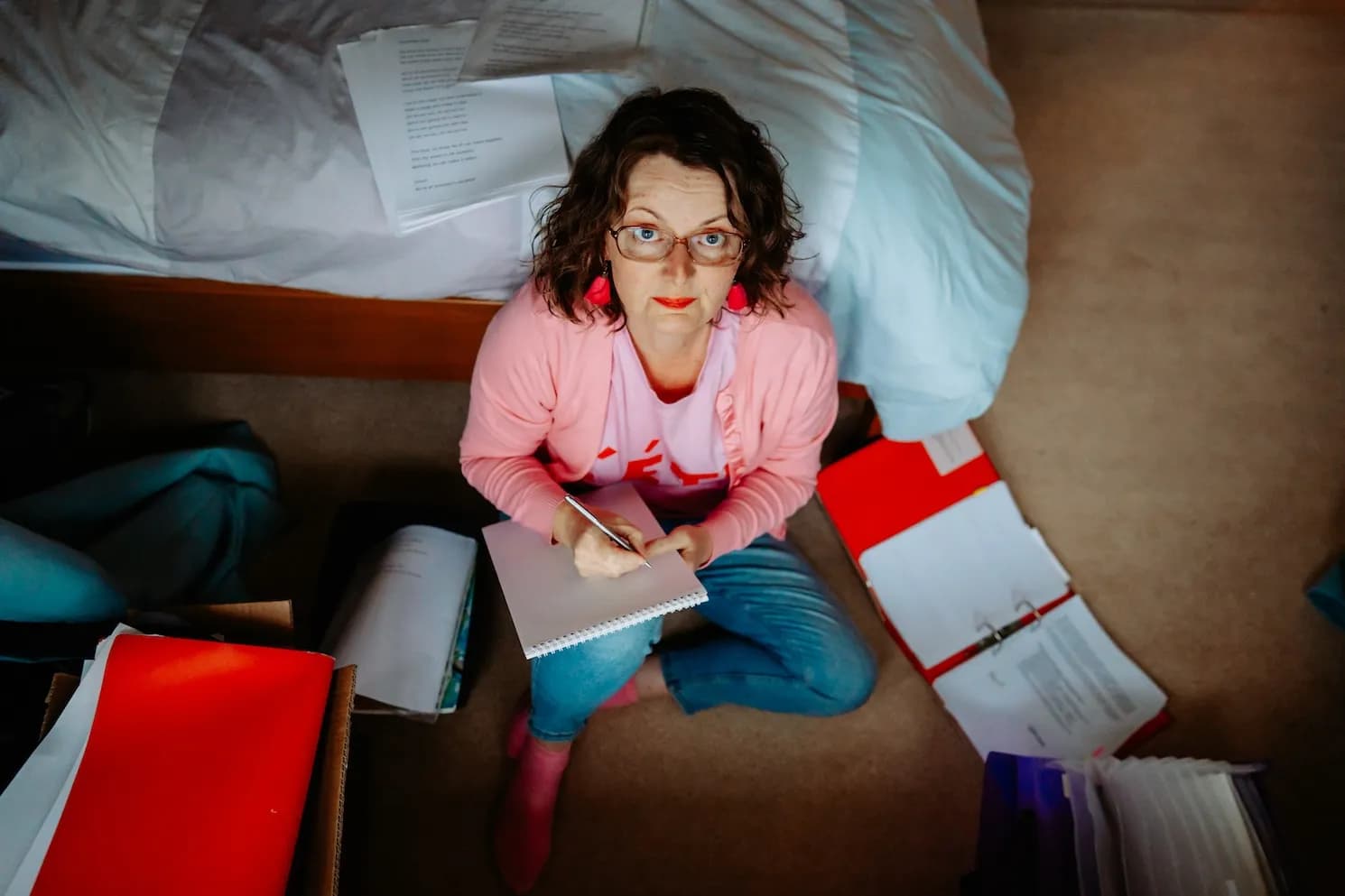 A woman with glasses and red earrings sits on the floor writing in a notebook, surrounded by papers and folders.