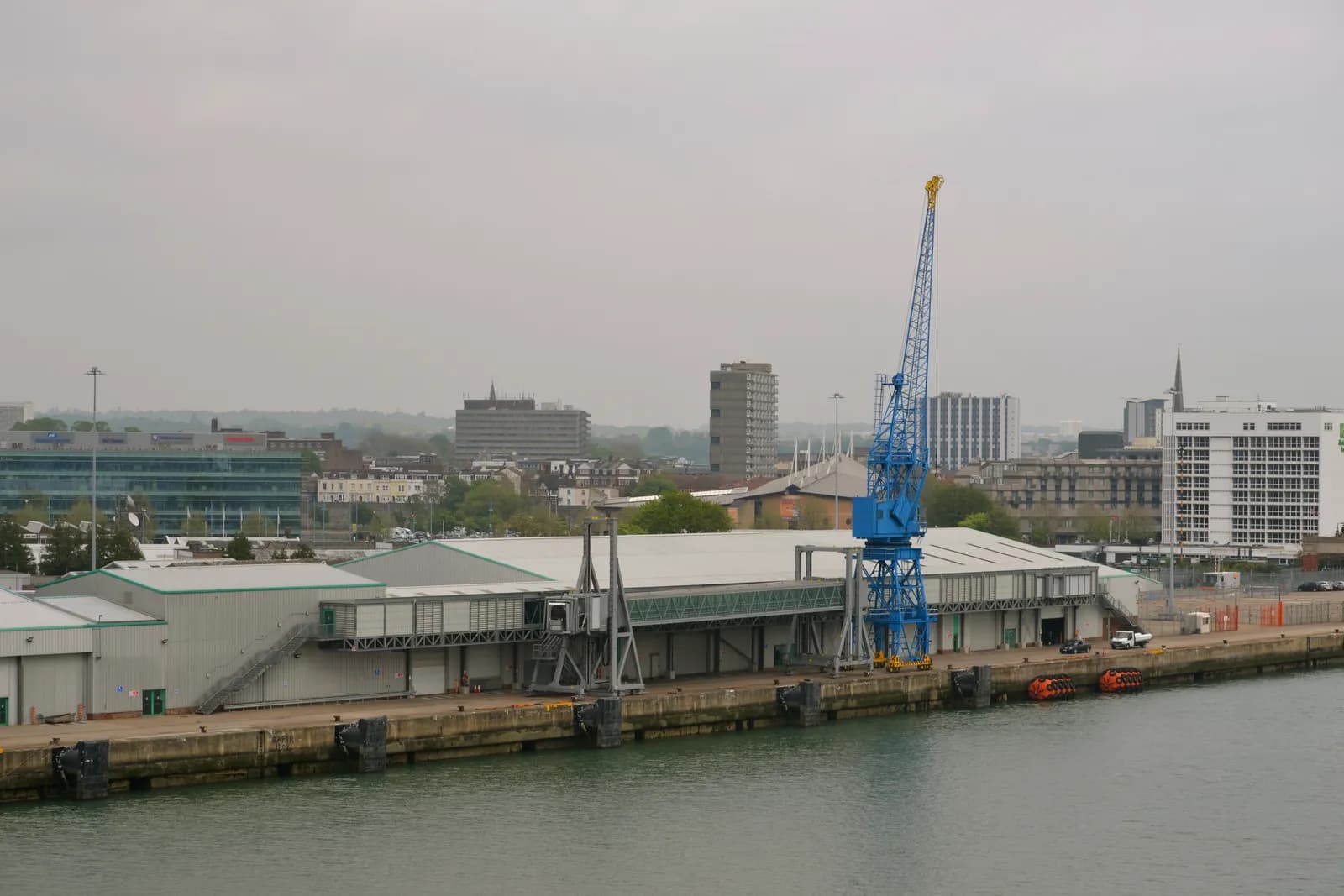 A large blue crane stands prominently on a dock next to warehouse buildings, with urban buildings and a hazy sky in the background.