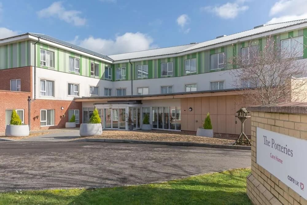 A modern, multi-story building with green and white striped upper floors and brick lower sections, featuring a prominent glass entrance and a sign for 'The Potteries Care Home' in the foreground.