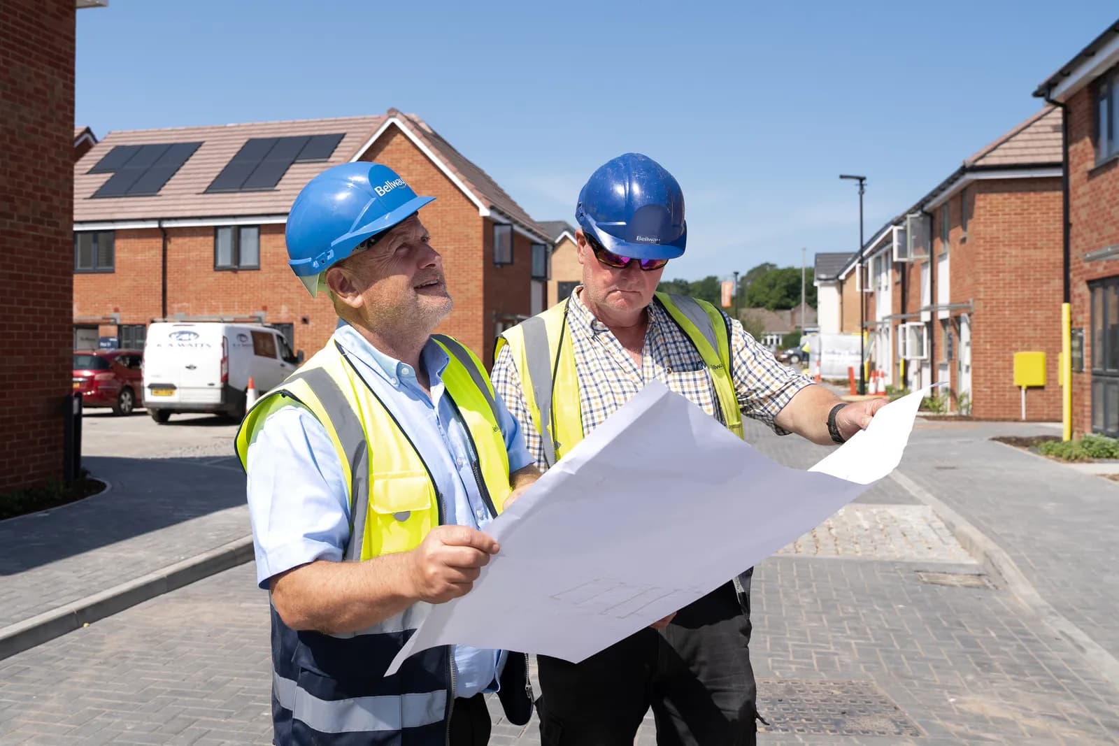Two male construction workers in hard hats and safety vests are examining blueprints on a paved road at a construction site with new brick buildings in the background.
