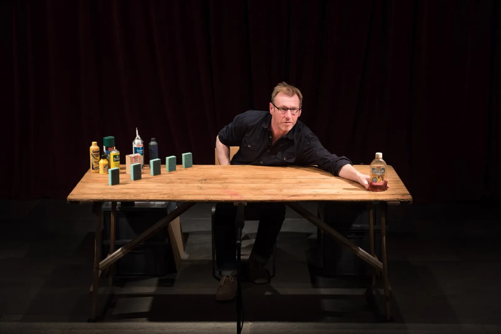 A man in a black shirt and glasses sits behind a wooden table with various cleaning supplies and small rectangular sponges arranged on it.