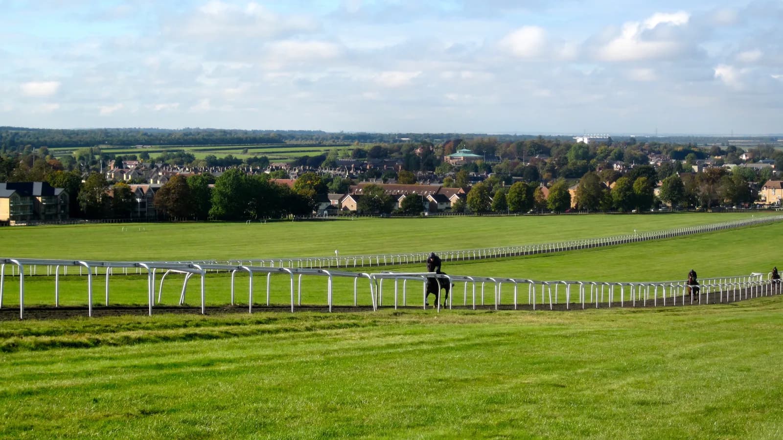 A vast green horse racing track stretches into the distance, with a white rail fence bordering the inner lane where a jockey on a dark horse is visible, and in the background, a town with houses and trees under a partly cloudy sky.