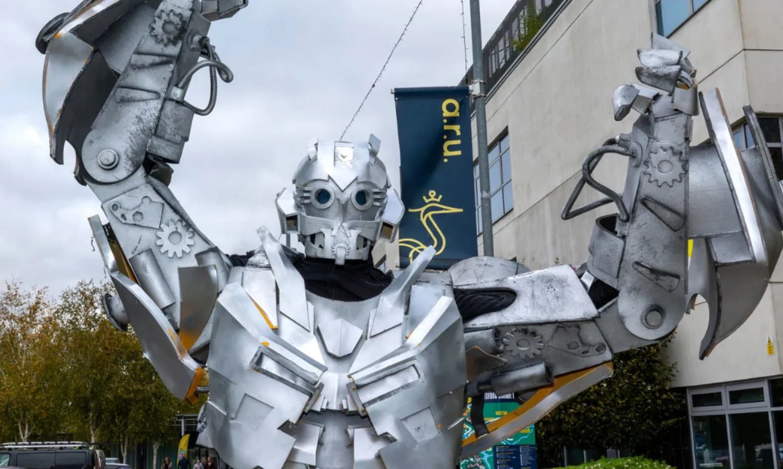A person in an intricate silver robot costume with arms raised stands outdoors in front of a building with a blue and yellow banner.