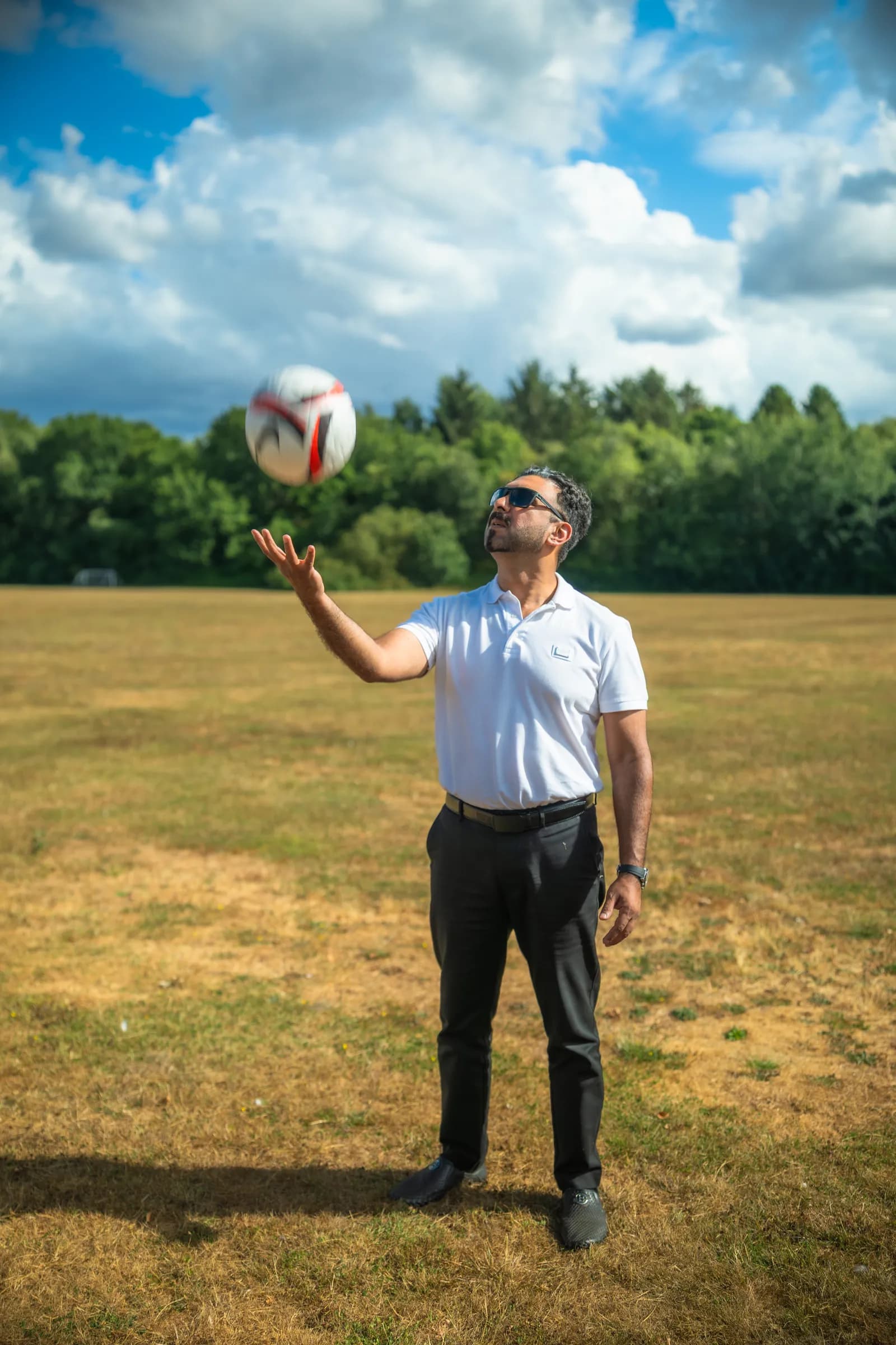 A man in a white polo shirt and sunglasses looking up as he bounces a white and red soccer ball on his left hand in a grassy field.