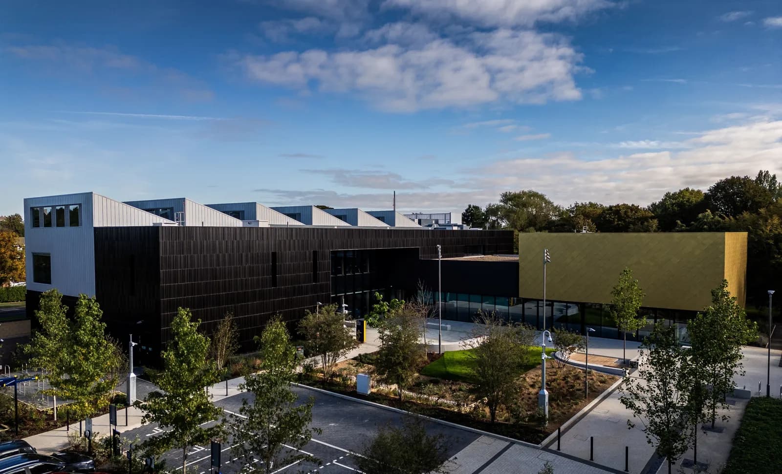 A modern building with a dark textured facade and a gold-colored section, under a partly cloudy sky, with trees and a parking lot in the foreground.