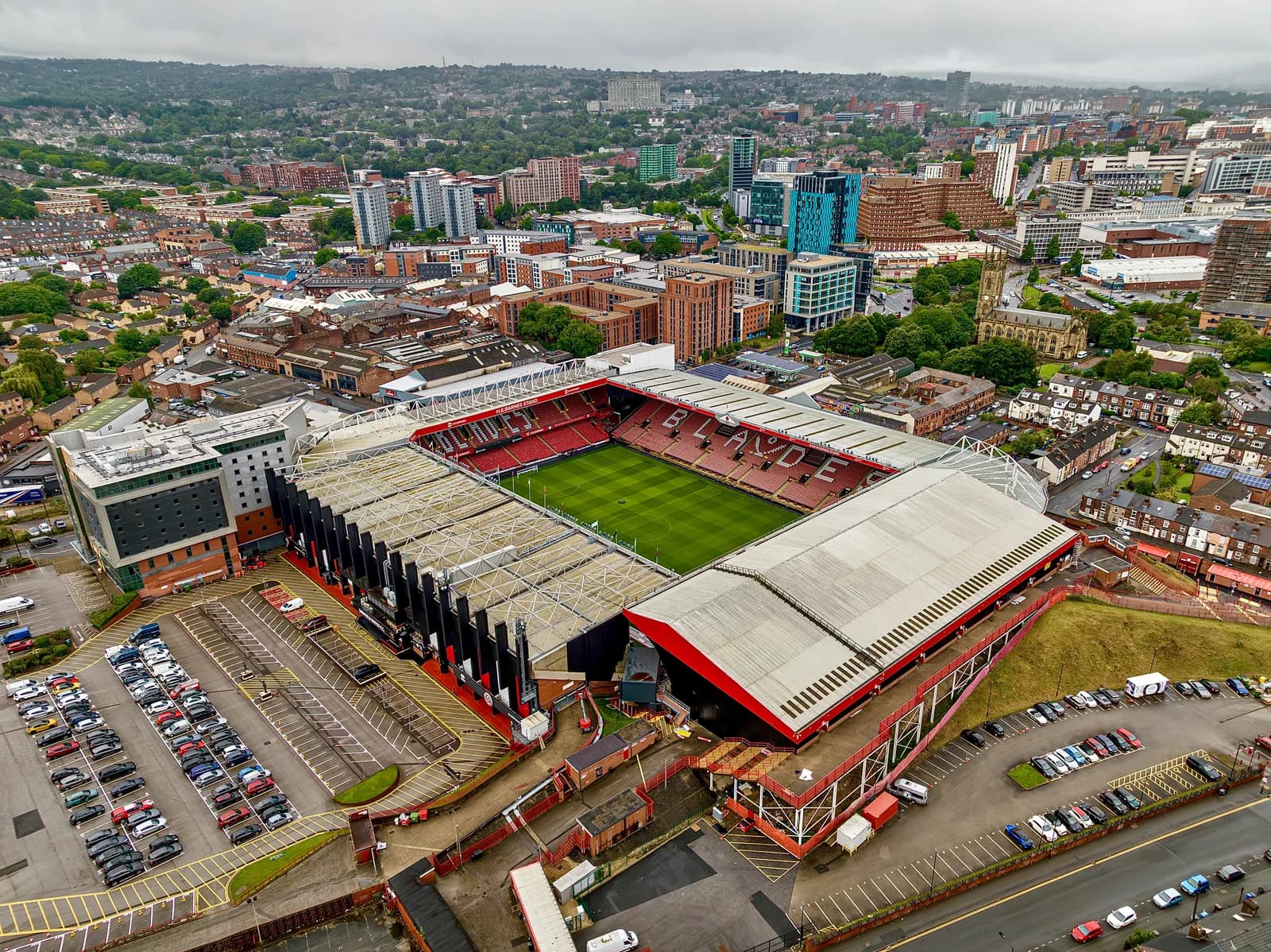 An aerial view of a large sports stadium with a bright green field, surrounded by parking lots and a densely built-up city with many buildings and some green hills in the distance under an overcast sky.