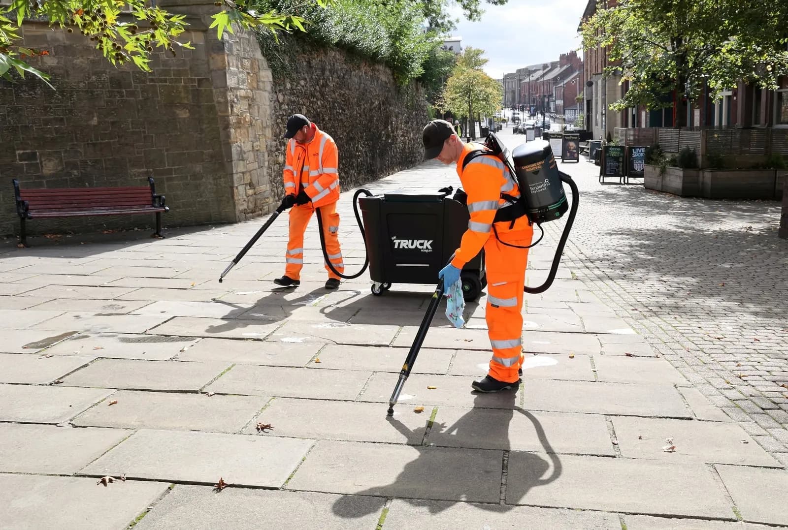 Two municipal workers in orange uniforms clean a stone-paved path with specialized vacuum-like devices, with one worker carrying a backpack unit and both connected to a central cleaning machine on wheels.
