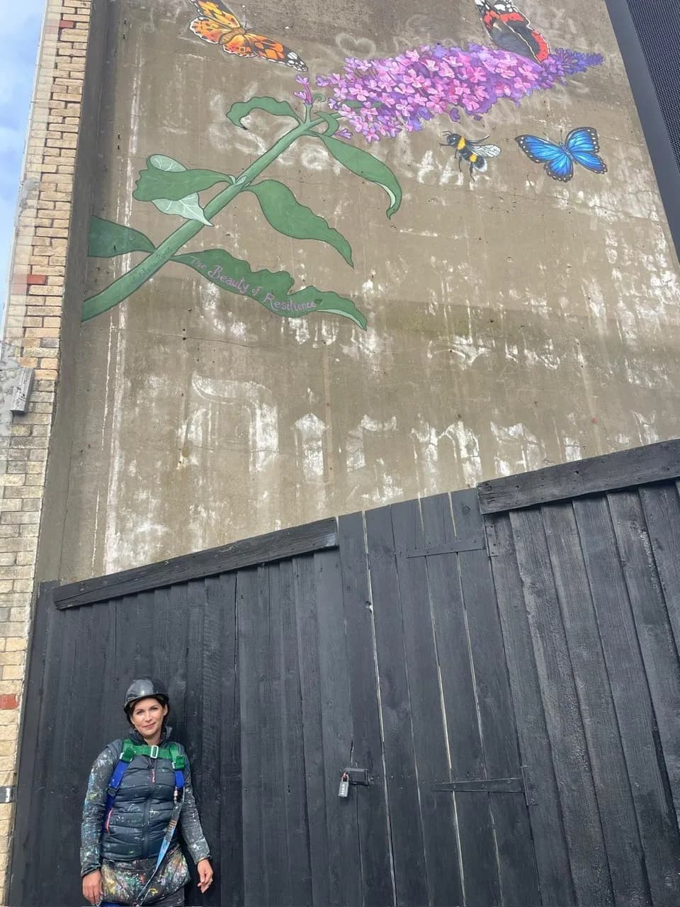 A woman wearing a helmet and painting harness stands in front of a tall wooden gate with a large mural of butterflies, a bee, and a purple flower on the concrete wall above her.