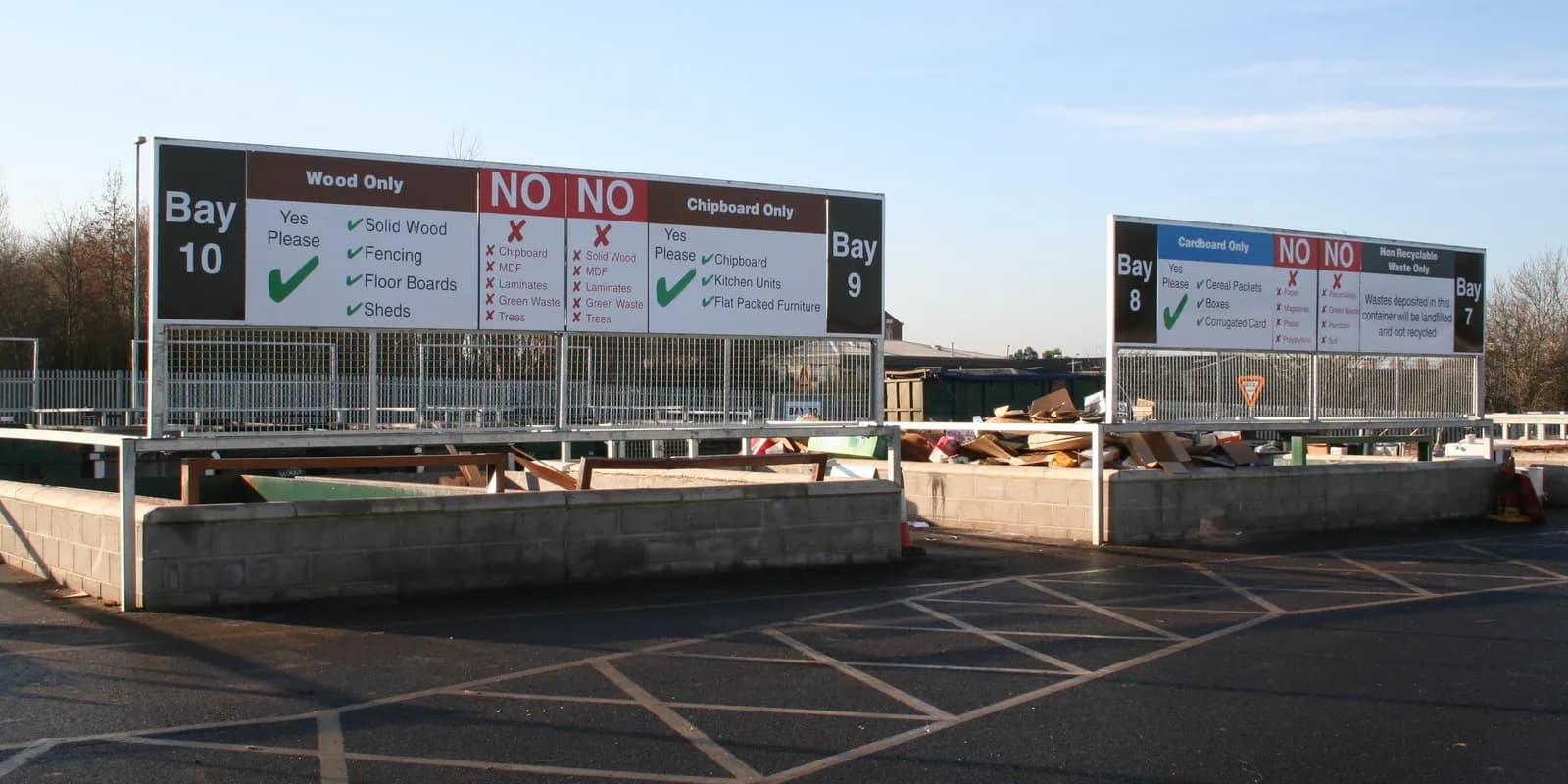 Several large white and brown signs at a recycling center with instructions for sorting different types of waste, including wood, chipboard, and cardboard.