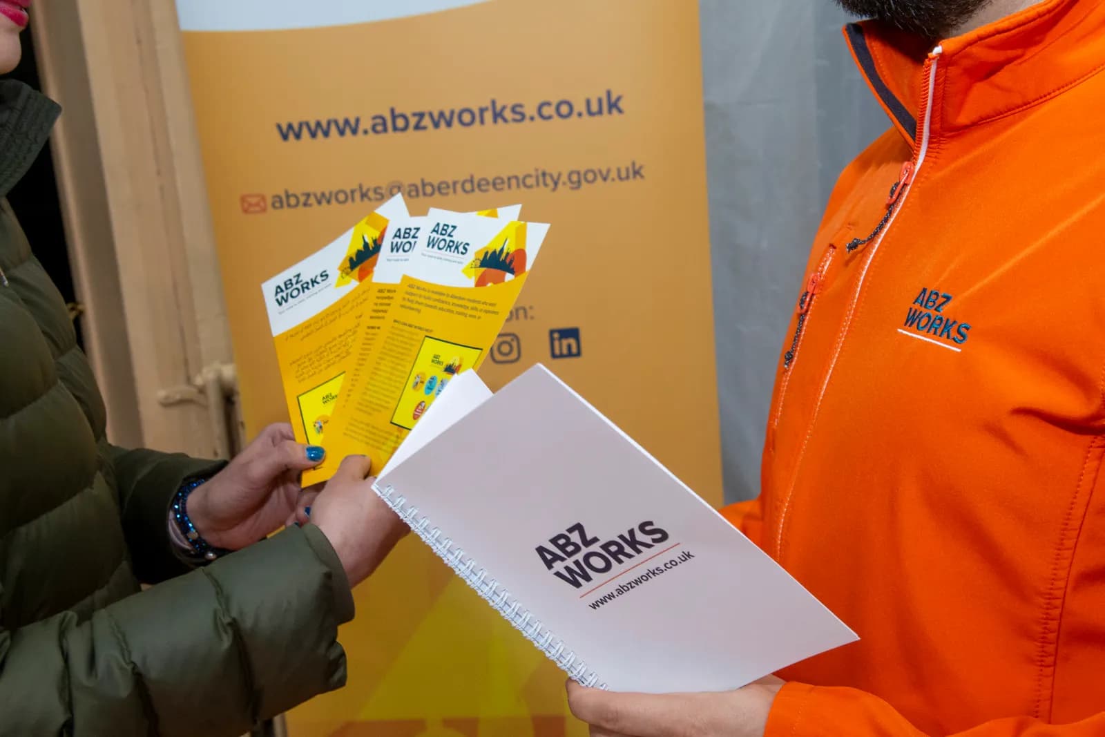 Two people exchanging promotional materials for Abzworks, including a brochure and a spiral-bound notebook, in front of a yellow banner.