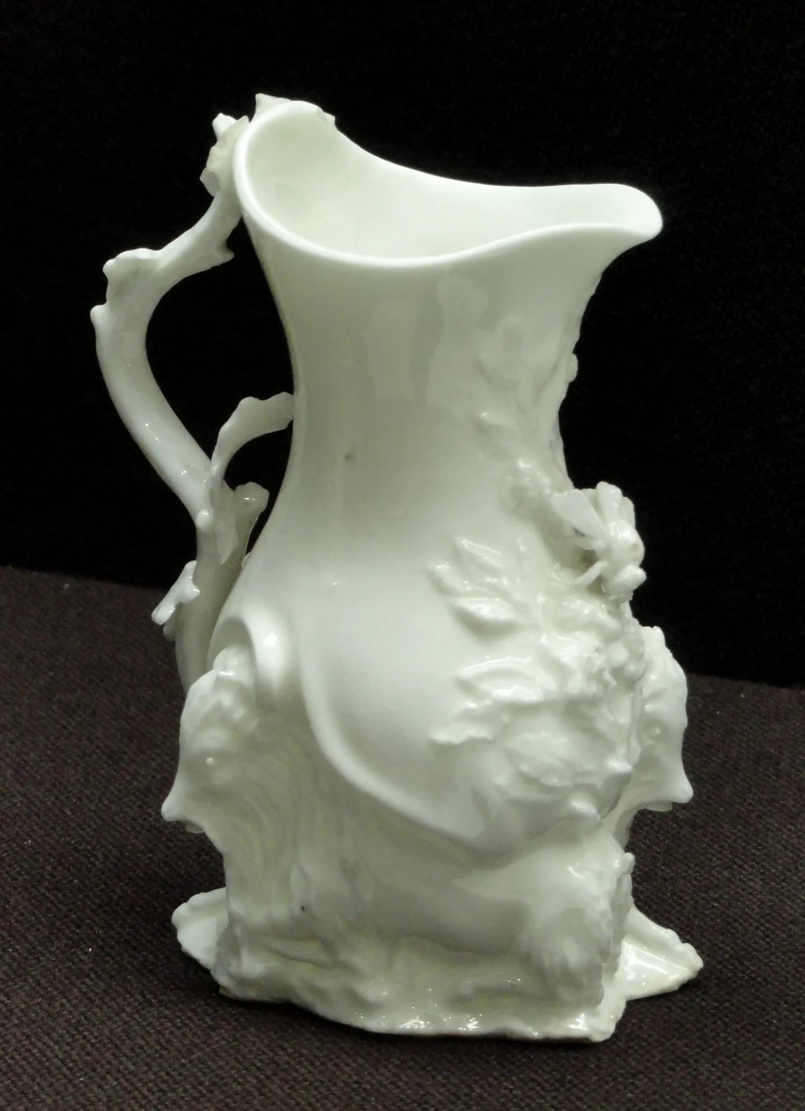 A white porcelain ewer with intricate raised floral and figural designs on its body and a branch-like handle.