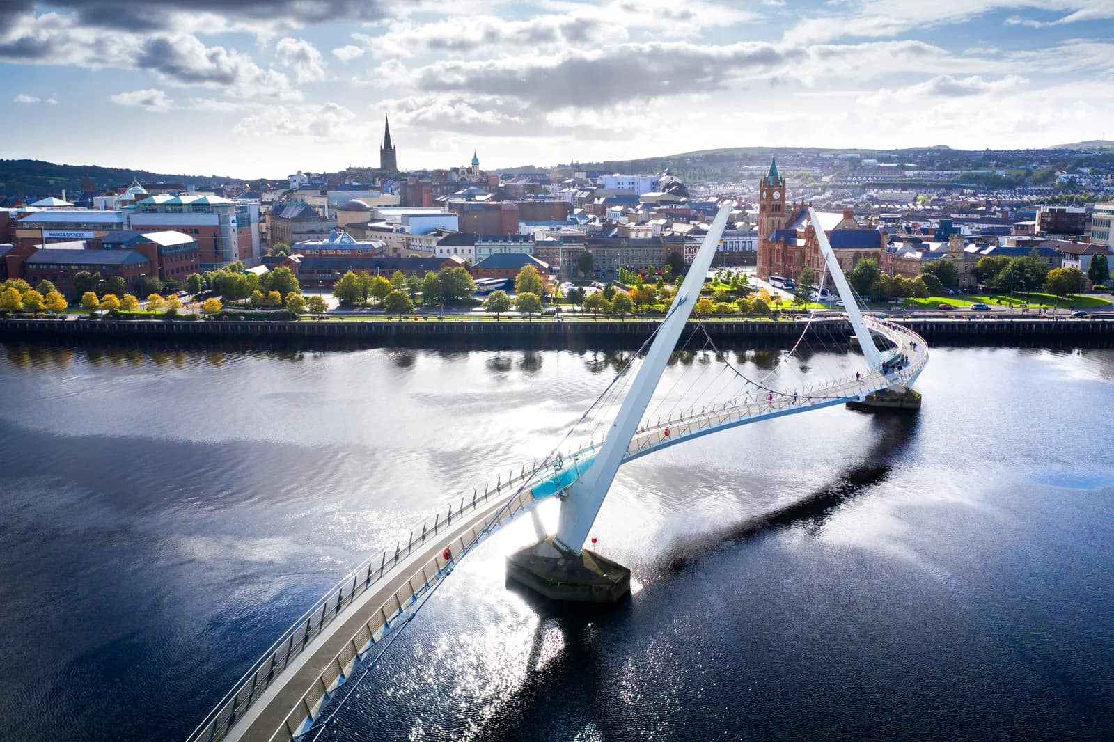 A wide aerial shot shows a modern white pedestrian bridge curving across a dark blue river towards a city skyline with various buildings and a tall church steeple under a partially cloudy sky.