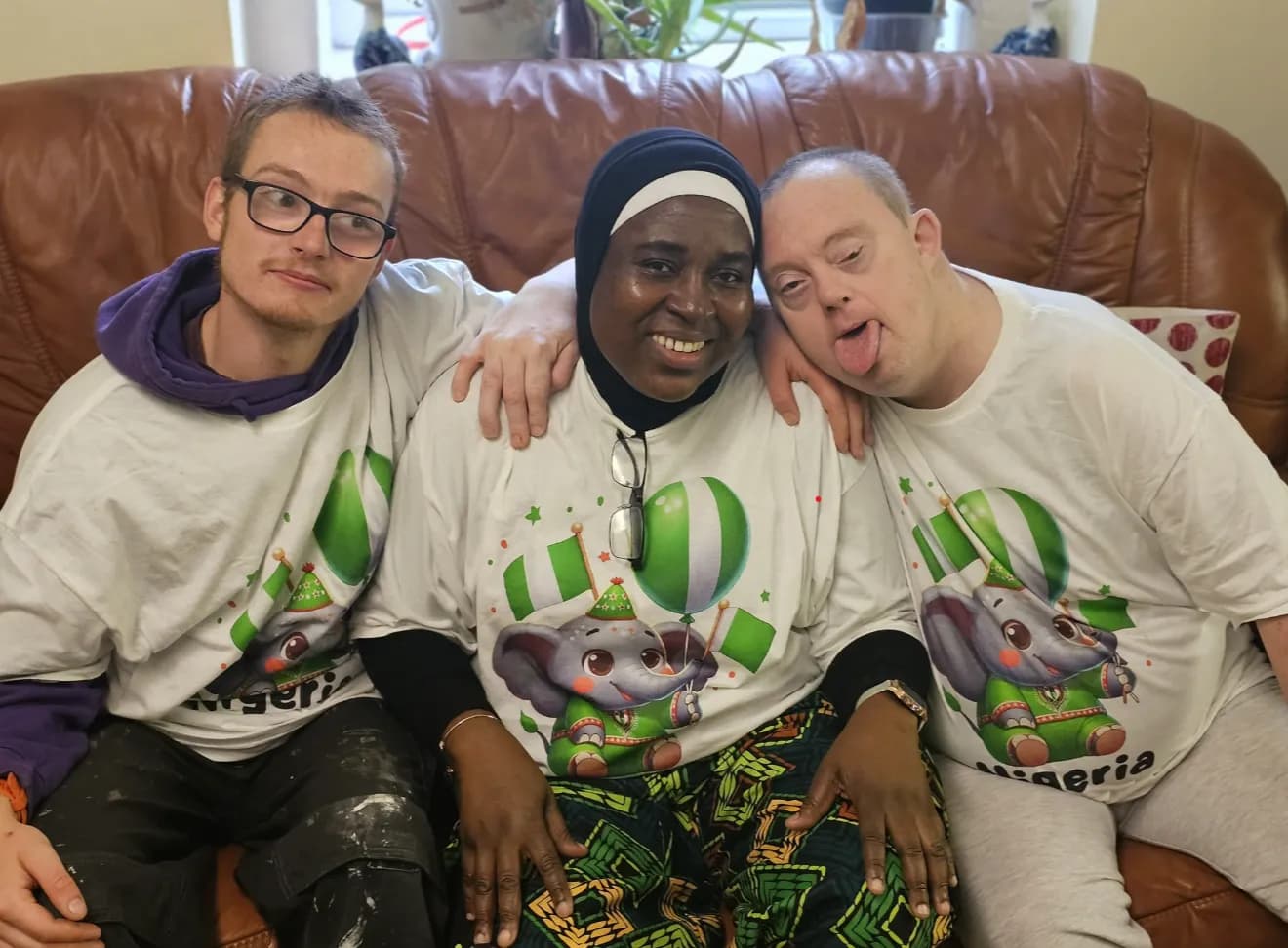 Three people, two men and one woman, sit together on a brown leather couch, smiling and wearing white t-shirts with a cartoon elephant holding green and white balloons and Nigerian flags.