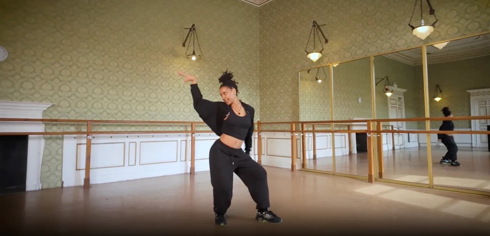 A woman in a black crop top and sweatpants dances in a studio with green patterned walls, a wooden barre, and large mirrors.